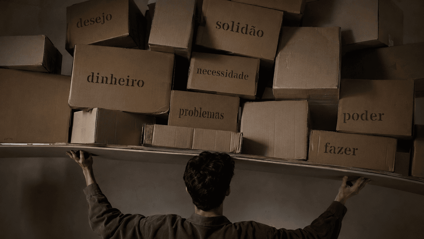 Homem de costas sustenta uma pilha irregular de caixas com palavras como desejo, solidão, dinheiro, poder, problemas, necessidade e fazer.