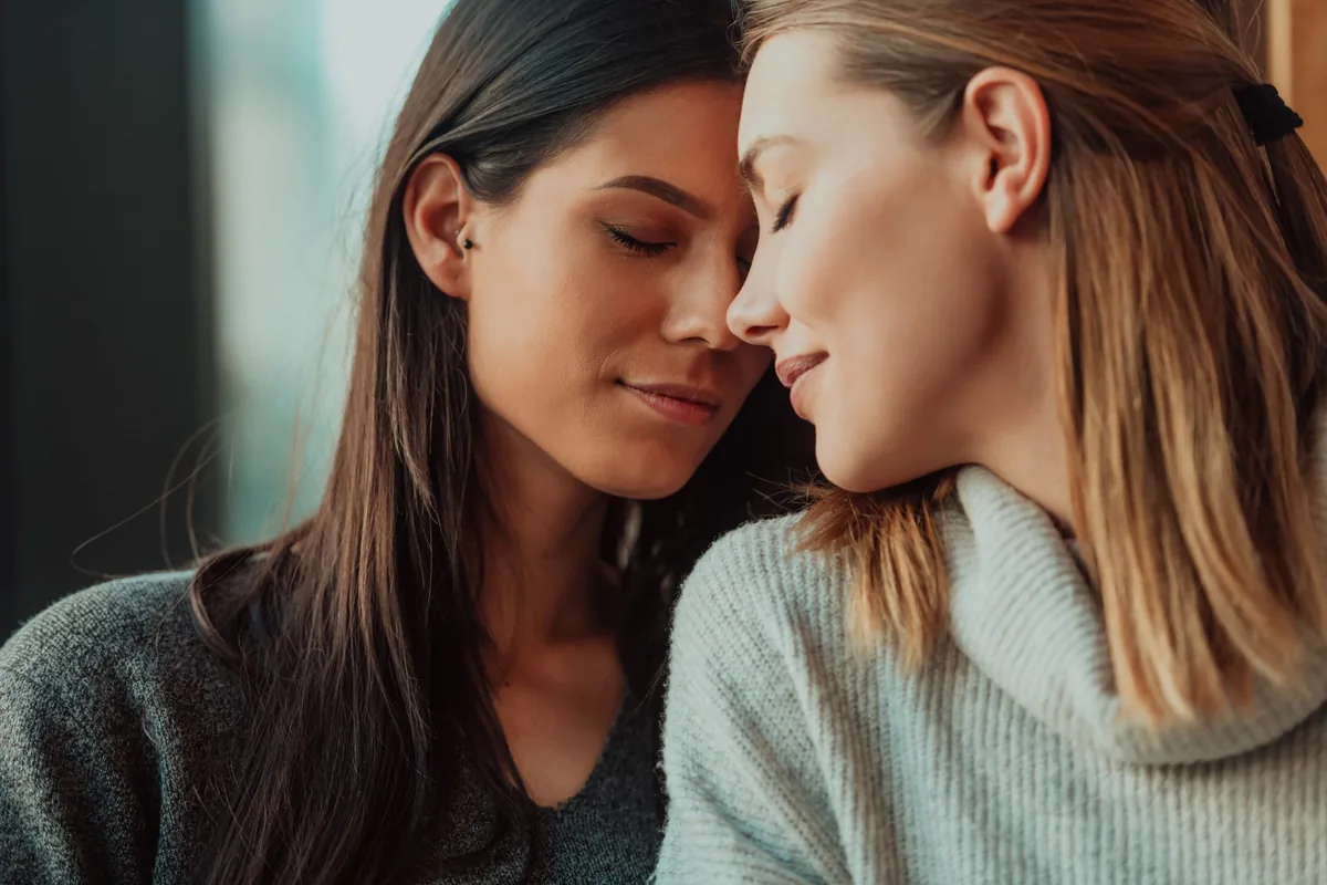 Duas mulheres adultas aproximam os rostos e fecham os olhos em um momento de intimidade e afeto.