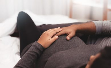 Close up of pregnant couple lying on bed at home. Focus on hands of man and woman on a pregnant belly.
