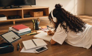 Adolescente com fones de ouvido desenhando no chão entre cadernos, tablet e materiais escolares.