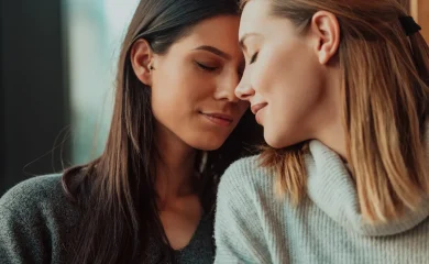 Duas mulheres adultas aproximam os rostos e fecham os olhos em um momento de intimidade e afeto.