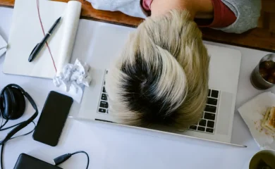 Pessoa exausta sobre o notebook em uma mesa com celular, fones de ouvido, papel amassado, refrigerante e resto de sanduíche, representando sobrecarga no TDAH em adultos.
