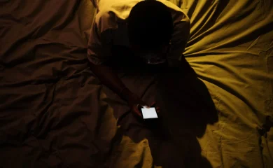 Homem sentado na cama usando o celular à noite, em artigo sobre uso problemático de pornografia.